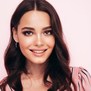 Fashion beauty portrait of young brunette woman with evening stylish  makeup and perfect clean skin. Smiling model with long curly hair posing in studio on pink background
